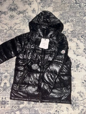 Svart dunjacka med huva från Moncler - Snygg svart dunjacka från Moncler med glansig finish och huva. Jackan har dragkedja framtill, två fickor med dragkedja och Moncler-logga på ärmen. Insidan har ett unikt serietidningsmönster och märkning. Perfekt för kalla vinterdagar.