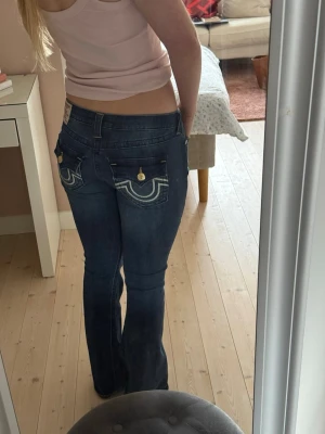 Blå bootcut jeans från True Religion - Säljer ett par mörkblå bootcut jeans från True Religion med jättesnygga kontrastsömmar och detaljerade fickor bak. Jeansen har låg midja, guldknappar och klassisk logga på bakfickan. Endast använda två gången så i nyskick! 