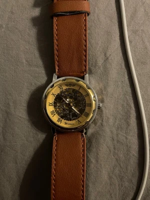 Guldig Winner klocka med brunt läderband - Stilren klocka från Winner med guldig urtavla och synlig mekanik. Klockan har romerska siffror och ett brunt läderband med kontrastsömmar. Boetten är i silverfärgad metall och baksidan har genomskinligt fönster som visar urverket. Perfekt accessoar för dig som gillar detaljer.