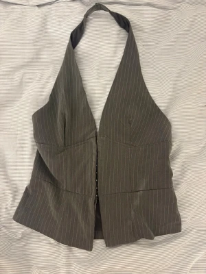 Grå randig halterneck väst - Snygg grå väst med smala vita ränder och halterneck-modell. Västen har djup v-ringning och knäppning framtill. Perfekt för att skapa en trendig och stilren look. Har sytt in den i midjan till en storlek s. 