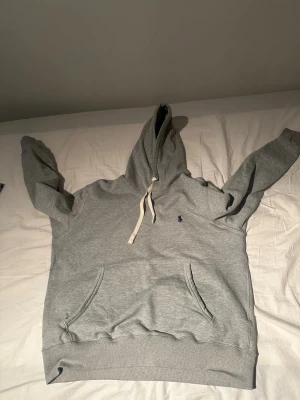 Grå hoodie från Polo Ralph Lauren - Snygg grå hoodie från Polo Ralph Lauren med klassisk känguruficka och vit dragsko i huvan. Liten mörkblå logga broderad på bröstet. Tillverkad i mjuk bomullsblandning som känns skön mot huden. Perfekt för en avslappnad och stilren look.pris går att diskutera.