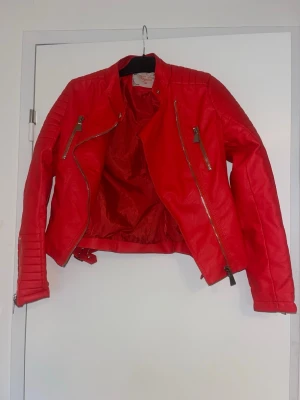 Röd biker skinnjacka med dragkedjor - Snygg röd skinnjacka i bikerstil med coola detaljer som quiltade partier på axlar och ärmar samt flera dragkedjor framtill och på ärmarna. Kort modell med bälte i nederkant och ståkrage med tryckknapp. Perfekt för dig som vill sticka ut. Den är i storlek 40