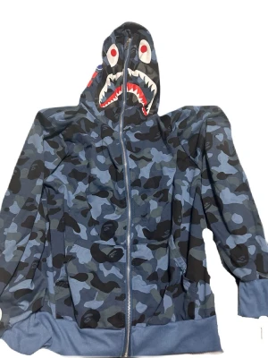 BAPE blå camo shark hoodie - Säljer en ikonisk BAPE shark hoodie i blå camo med dragkedja hela vägen upp i huvan. Huvan har hajmotiv med tänder och ögon framtill samt broderade bokstäver och tiger på sidan. Tillverkad i mjuk bomullsmix och har ribbade muddar. (Inte äkta!), storlek M-L