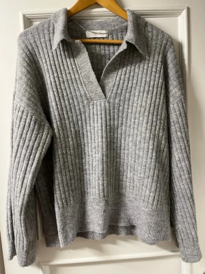 Grå ribbstickad tröja Samsa - Säljer en grå ribbstickad tröja från Samsa Collection. Tröjan har en avslappnad passform, krage och djup v-ringning. Materialet är mjukt och passar perfekt för kyligare dagar. Långa ärmar och breda muddar ger en trendig look.