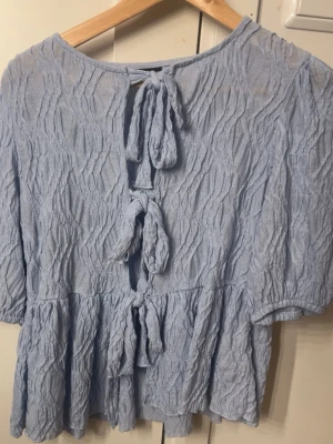 Ljusblå blus med knyt och struktur - Supersöt ljusblå blus med tre knyt i ryggen och ett vågigt, strukturerat tyg. Blusen har korta ärmar och volang nedtill, perfekt för en trendig och luftig look.