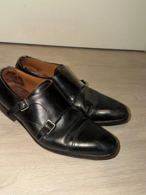Svarta dubbla monkstrap loafers - Snygga svarta loafers med dubbla spännen i metall och klassisk cap-toe design. Skorna har en elegant, smal siluett och är tillverkade i slätt läder med brunt innerfoder. Perfekta för en stilren och dressad look.