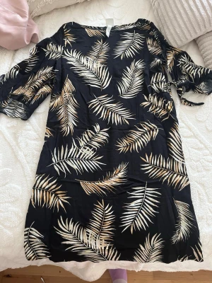 Svart kortklänning med palmblad H&M - Svart kortklänning från H&M med beige och vita palmbladsmönster. Klänningen har korta ärmar med knytband och en avslappnad passform. Perfekt för sommarens varma dagar och enkel att styla med sneakers eller sandaler.