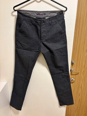 Mörkgrå chinos med smal passform - Snygga mörkgrå chinos med smal passform och klassisk design. Byxorna har snedställda fickor fram, två bakfickor och bälteshällor. Insidan av linningen har ett randigt tyg för extra detalj. Perfekta för en stilren look.