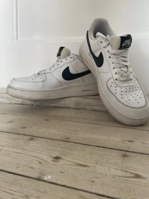 Nike Air Force 1 vita sneakers - Klassiska vita Nike Air Force 1 sneakers med svart swoosh och detaljer. Lågt skaft, perforerad tå och robust sula. Skön passform och tidlös design som funkar till det mesta. Tillverkade i läder med textilfoder och gummisula.