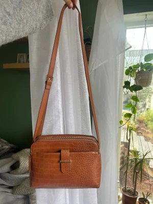 Brun axelväska i krokopräglat skinn - Snygg brun axelväska i krokopräglat konstläder med justerbar rem och guldfärgad detalj på framsidan. Väskan har dragkedja upptill och ett stilrent, fyrkantigt format som passar till många outfits. Perfekt för dig som gillar trendiga accessoarer.