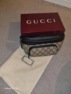 Gucci axelväska med GG-mönster - Snygg axelväska från Gucci i beige canvas med klassiskt GG-monogram och svarta läderdetaljer. Väskan har två dragkedjefack och justerbar rem. Perfekt för dig som vill ha en trendig och praktisk väska med lyxig känsla.