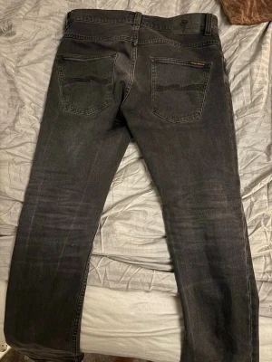 Svarta jeans från Nudie Jeans - Säljer ett par svarta jeans från Nudie Jeans med klassisk femficksmodell och raka ben. Jeansen har subtila slitningar och Nudie-loggan på bakfickan. Tillverkade i bomull med snygg tvättad finish. Perfekta för en avslappnad och stilren look.