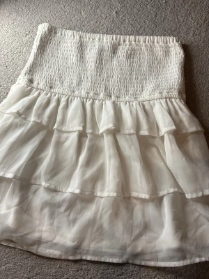 Vit volangkjol från BikBok - Supersöt vit kortkjol från BikBok med smock i midjan och flera lager volanger. Kjolen är gjord i ett lätt och luftigt material som ger en flowy känsla. Perfekt för sommarens alla tillfällen och enkel att styla med crop tops eller linnen. Hör av dig vid frågor!✨🥰