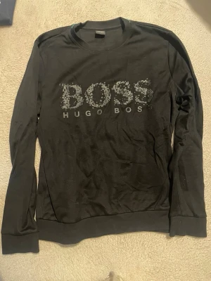 Svart långärmad tröja Hugo Boss - Svart långärmad tröja från Hugo Boss med stort BOSS-tryck på bröstet i grått och vitt. Rund halsringning och ribbade muddar vid ärmslut och nederkant. Perfekt för en stilren look och enkel att matcha med jeans eller joggers.