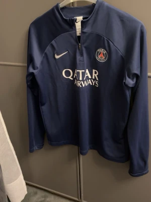 Mörkblå PSG träningströja Nike - Snygg mörkblå träningströja från Nike med Paris Saint-Germain-logga och Qatar Airways-tryck på bröstet. Tröjan har halv dragkedja framtill och är tillverkad i Dri-FIT-material som andas, perfekt för fotbollsträning eller sport.