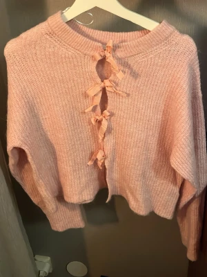 Rosa stickad tröja med rosetter - Supersöt rosa stickad tröja med unika rosettdetaljer längs framsidan. Tröjan har en lös passform, långa ärmar och ribbade muddar. Materialet ser mjukt och fluffigt ut, perfekt för att hålla värmen. 