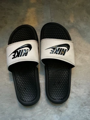 Svarta och vita Nike tofflor - Svarta och vita Nike tofflor i storlek 38,5 men passar mig som har 39/40. Otroligt bekväm sula och perfekta för sommaren. Inga skavanker utom lite smuts.