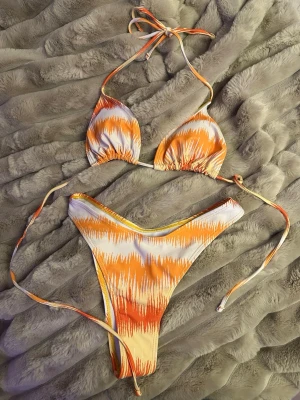 Orange och vit bikini med knyt - Trendig bikini i orange och vitt med coolt mönster och smala knytband. Överdelen är trekantsmodell och underdelen har hög skärning. Perfekt för strandhäng och soliga dagar.