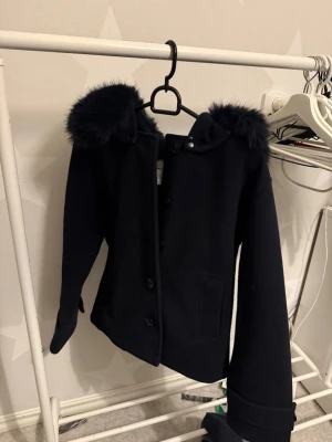 Svart kappa med fuskpäls - Säljer en svart kappa med huva i storlek 170 då den inte används. Använd ca 2 gånger sedan i höstas. Huvan går att ta av med knappar om man vill det. ☺️ Passar inte priset så lägg ett prisförslag. Nypris 499kr