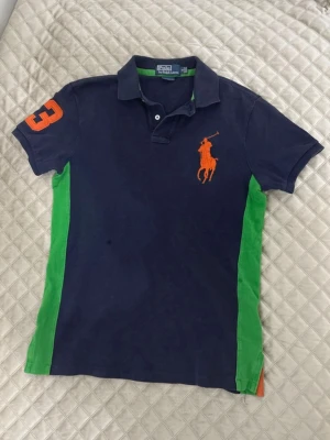 Mörkblå pikétröja från Ralph Lauren - Snygg mörkblå pikétröja från Ralph Lauren med gröna paneler på sidorna och orange logga på bröstet. Orange siffra på ärmen och klassisk krage med knappar. Perfekt för en sportig och avslappnad look.