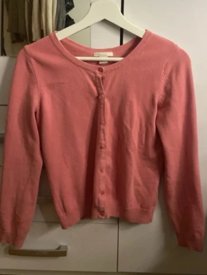 Rosa kofta med rund hals - Söt rosa kofta med rund halsringning och knappar framtill. Långärmad modell i mjukt, tunt material som passar perfekt till lager på lager. Enkel och stilren design utan mönster eller detaljer.