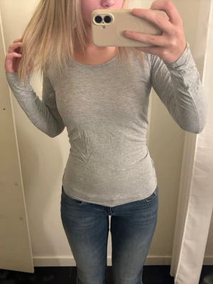 Ljusgrå långärmad topp - En enkel och stilren ljusgrå långärmad topp i mjukt material. Modellen är figurnära med rund halsringning och passar perfekt till jeans eller kjol. Toppen har en clean look utan synliga detaljer eller mönster.