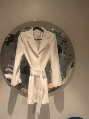 Vit  kavaj klänning med bälte - Säljer en stilren vit klänning från Nelly med lång ärm och snygg krage. Klänningen har en figurnära passform och medföljande bälte som markerar midjan. Perfekt för dig som gillar minimalistisk design och vill ha något elegant.perfekt till student osv 