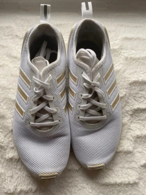 Vita Adidas sneakers med beige ränder - Snygga vita sneakers från Adidas med klassiska tre beige ränder på sidorna. Skorna har ovandel i meshmaterial för bra andningsförmåga och vita snören. Perfekta för en sportig och avslappnad stil.