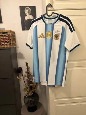 Argentina fotbollströja Adidas (authentic) - Snygg Argentina fotbollströja från Adidas med klassiska ljusblå och vita ränder, guldiga detaljer och AFA-emblem på bröstet. Tröjan har korta ärmar, rund hals och tre mörkblå ränder på axlarna. Perfekt för fotbollsfans!