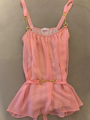 Rosa linnetopp med gulddetaljer - Säljer en söt rosa linnetopp från Ida sjödstedt  med transparent tyg, guldfärgade metalldetaljer på axelbanden och ett dekorativt bälte i midjan. Toppen har volangkant nedtill och justerbara axelband för perfekt passform.