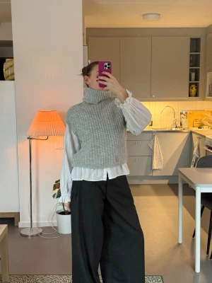 Grå stickad väst med polokrage - Snygg grå stickad väst med hög polokrage och ribbad struktur. Perfekt att bära över en skjorta för en trendig lager-på-lager-look. Västen har en croppad passform och är riktigt mjuk och skön.