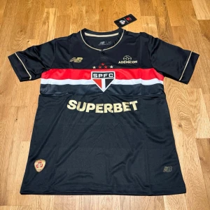 São Paulo away kit 25/26  - Snygg svart matchtröja från São Paulo FC med guldiga detaljer och logotyper. Tröjan har röda och vita ränder över bröstet, klubbmärke och sponsortryck. Tillverkad i lätt och ventilerande funktionsmaterial, perfekt för fotboll eller träning.