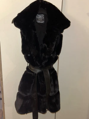 Svart pälsväst från Amisu med bälte - Snygg svart pälsväst från Amisu i mjuk fuskpäls. Västen har en bred krage och knyts i midjan med ett matchande svart bälte i läderimitation. Perfekt för att lyfta din outfit med en lyxig touch och extra värme.