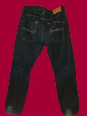 Nudie Jeans - Säljer ett par mörkblå jeans från Nudie Jeans 2001 med klassisk femficksmodell och kontrastsömmar. Jeansen har raka ben, snygga slitningar framtill och broderade bakfickor. Tillverkade i kraftigt denimtyg som ger en cool och tidlös look.