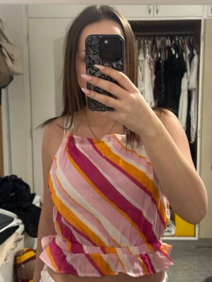 Randig linnetopp med volang - Säljer en färgglad randig linnetopp med smala axelband och volang nertill. Toppen har diagonala ränder i rosa, gul, orange och vit, och är tillverkad i bomull. Perfekt för sommarens varma dagar och ger en lekfull vibe.