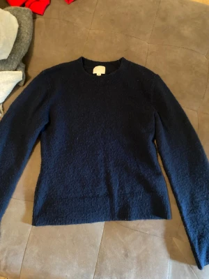 Mörkblå stickad tröja från H&M - Mysig mörkblå stickad tröja från H&M med rund halsringning och långa ärmar. Tröjan har en enkel, klassisk design och är tillverkad i ett mjukt material som passar perfekt för kyligare dagar.