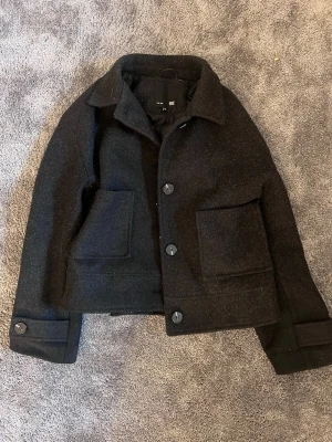 Svart kappa  - Stilren svart kappa med klassisk krage, stora framfickor och knäppning med tre knappar. Rocken har en rak passform och detaljerade manschetter med knapp. Perfekt för dig som gillar minimalistisk och tidlös design.