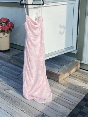 Råda Balklänning  - Säljer en ljusrosa lång balklänning med tunna axelband och diskret blommigt mönster. Klänningen har en avslappnad passform och mjukt, glansigt tyg som ger en lyxig känsla. Perfekt för dig som gillar romantisk och trendig stil.