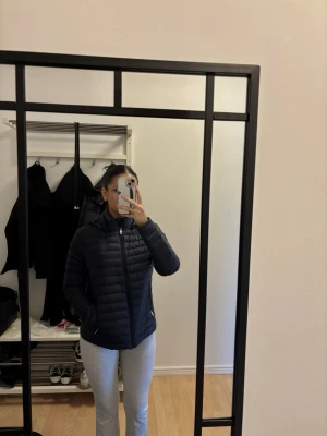 Svart pufferjacka med huva - Säljer en svart pufferjacka med huva och dragkedja framtill. Jackan har en smal passform och horisontella sömmar som ger en klassisk pufferlook. Perfekt för kalla vinterdagar och enkel att matcha med olika outfits.