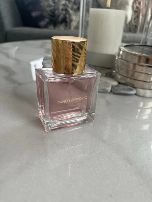 Panos Emporio parfym rosa glasflaska - Elegant parfym från Panos Emporio i en stilren, fyrkantig glasflaska med rosa innehåll och lyxig guldfärgad kork. Perfekt för dig som vill ha en fräsch och modern doft i snygg design.