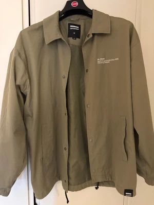 Olivgrön vindjacka från Dr Denim - Stilren olivgrön vindjacka från Dr Denim med tryckknappar, dragsko i nederkant och två sidofickor. Jackan har klassisk krage och diskret vit text på bröstet. Tillverkad i ett lätt material som passar perfekt till streetwear och layering.