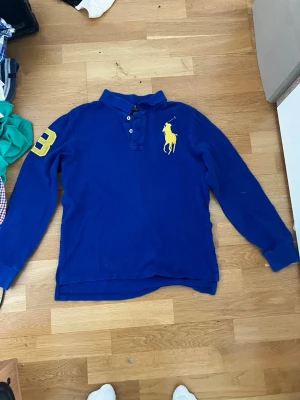 Blå långärmad henley från Ralph Lauren - Blå långärmad henley från Ralph Lauren med stor gul logga på bröstet och siffran 3 i gult på ena ärmen. Skjortan har knappar vid halsen och är gjord i mjuk bomull. 