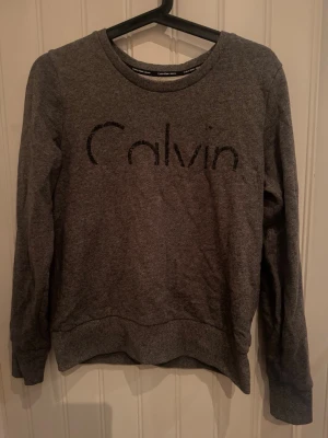 Grå sweatshirt från Calvin Klein Jeans - Mysig grå sweatshirt från Calvin Klein Jeans med rund halsringning och diskret logga framtill. Tröjan har långa ärmar och ribbade muddar vid ärmslut och nederkant. Perfekt för en avslappnad och stilren look.