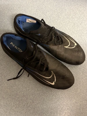 Nike Phantom svart/blå fotbollsskor - Säljer ett par Nike Phantom fotbollsskor i svart med blå detaljer och vit Nike-logga. Skorna har snörning och en låg profil, samt greppvänlig sula för bra fäste på planen. Insidan är blå och materialet är syntetiskt för lätt vikt och bra bollkontroll.Dom är storlek 44.5 passar även 44