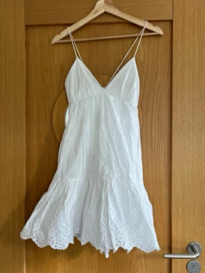 Vit sommarklänning med broderier - Söt vit klänning med tunna axelband och v-ringning. Klänningen har broderade detaljer och volangkant nertill, vilket ger en luftig och romantisk känsla. Perfekt för varma dagar och har en lätt, flowy passform.