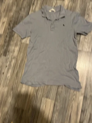 Ljusblå pikétröja från H&M - En ljusblå pikétröja från H&M med klassisk krage och knappslå framtill. Tröjan har korta ärmar och en liten broderad detalj på bröstet. Perfekt för en avslappnad och stilren look. Tillverkad i mjukt bomullsmaterial.