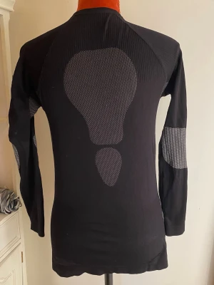Svart funktionsundertröja med mönster - Säljer en svart långärmad funktionsundertröja med grått meshmönster på ryggen och armbågarna. Tröjan är tight i modellen och har rund hals. Perfekt för träning eller som baslager vid utomhusaktiviteter.