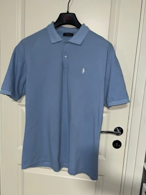 Blå pikétröja från Ralph Lauren - Klassisk ljusblå pikétröja från Ralph Lauren med vit broderad logga på bröstet. Tröjan har krage, knappar framtill och korta ärmar. Tillverkad i mjuk bomull som känns skön mot huden. Perfekt till jeans eller shorts.