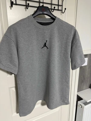 Grå Jordan t-shirt med logga - Snygg grå t-shirt från Jordan med den klassiska svarta Jumpman-loggan broderad på bröstet. T-shirten har rund hals, korta ärmar och är gjord i mjuk bomull för en skön känsla. Perfekt till streetwear-looken.