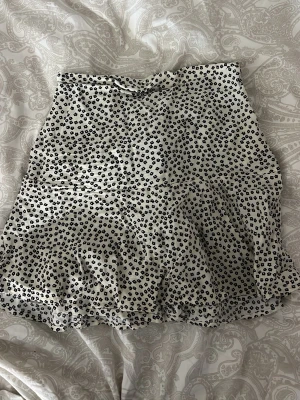 Blommig kortkjol från Zara - Supersöt kortkjol från Zara med svart blommönster på vit botten. Kjolen har en lätt volang nedtill och är perfekt för dig som gillar en feminin och trendig stil. Tillverkad i ett mjukt och skönt material som känns luftigt.